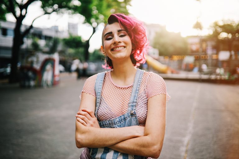 Women with bright pink hair standing in the street with arms crossed. Large smile and wearing a light pink shirt and denim overalls.